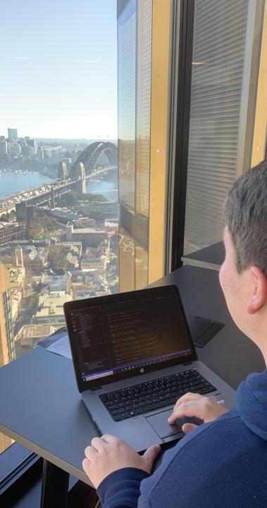 a student intern works on their computer while looking out the window at the Sydney Bridge