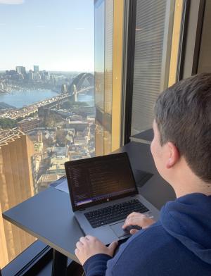 a student intern works on their computer while looking out the window at the Sydney Bridge