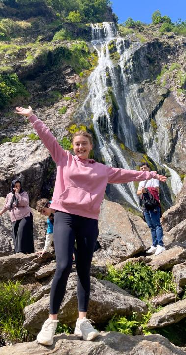 student standing on top of rock with outstretched hands in front of rocky grassy backdrop with a waterfall