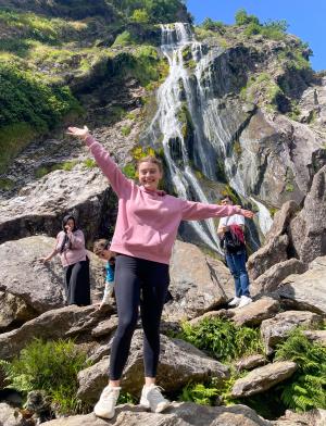 student standing on top of rock with outstretched hands in front of rocky grassy backdrop with a waterfall