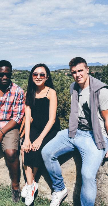 Three friends sitting on a short stone wall at a grassy park with an expansive background of trees behind them and mountains in the distance
