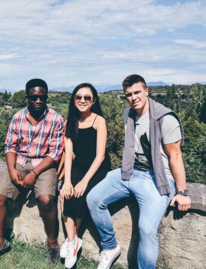 Three friends sitting on a short stone wall at a grassy park with an expansive background of trees behind them and mountains in the distance